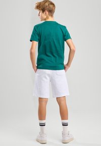 T-shirt verde in cotone, pantaloni corti bianchi in cotone e sneakers bianche con accenti neri. Modello di spalle, che mostra un outfit casual e in forma.
