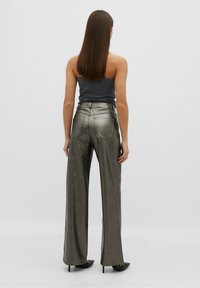 Pantalons larges métalliques verts avec une texture lisse, taille haute et poches arrière. Le modèle porte un haut ajusté de couleur sombre et des talons hauts.