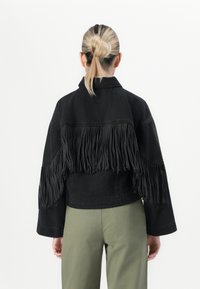Persona con capelli biondi raccolti in un bun, che indossa una giacca nera con frange e pantaloni verde oliva, mostrata di spalle su uno sfondo bianco.