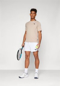 Nuori mies vaaleassa t-paidassa ja valkoisissa shortseissa, joka pitää tennismailaa yhdessä kädessä ja tennispalloa toisessa, seisoo yksinkertaista taustaa vasten.