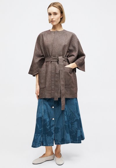 Γυναίκα με καφέ παλτό με ζώνη και φαρδιές τσέπες, μπλε τζιν παντελόνι καμπάνα με κουμπιά και μπεζ φλατ παπούτσια σε απλό φόντο.