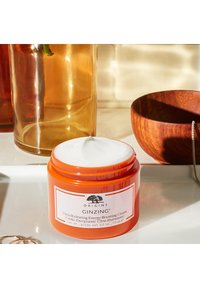 Oranssi muovipurkki, jossa on valkoinen kansi, etiketti "Origins Ginzing Ultra-Hydrating Energy-Boosting Cream," 75 ml. Ympärillä lasisia ja puukappaleita.