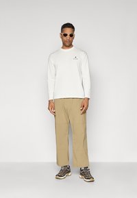 White long-sleeve shirt with small black logo, beige wide-leg pants, and grey sneakers with black accents. Model wears light-colored sunglasses.