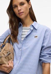 Blå og hvit rutete skjorte med avslappet passform, med en liten lapp, lagd over en grå og hvit stripete topp. Bærer en beige veske.