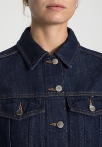 Dark blue denim jacket with a classic collar, silver buttons, chest pockets, and contrasting orange stitching. Smooth texture, structured design.