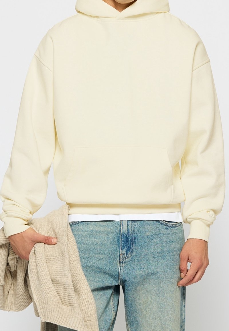 Personne portant un sweat à capuche jaune clair et un jean bleu, tenant un pull beige tricoté plié dans sa main gauche.