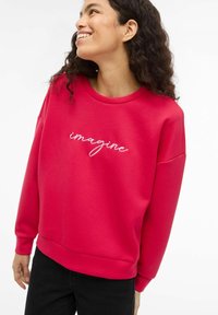 Červená mikina s kulatým výstřihem a klesajícími rameny. Na přední straně je bílý vyšitý nápis "imagine". Měkká texturovaná tkanina.