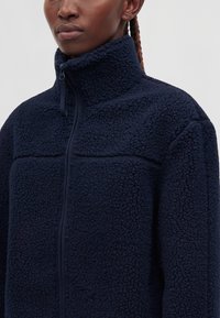 Personne portant une veste polaire bleu marine avec un col montant et une fermeture éclair frontale, montrant un tissu texturé et des cheveux tressés visibles derrière l'oreille.