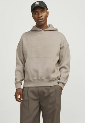 Beige hooded sweatshirt made of soft fabric, featuring a front pocket and ribbed cuffs, worn with tailored brown trousers.