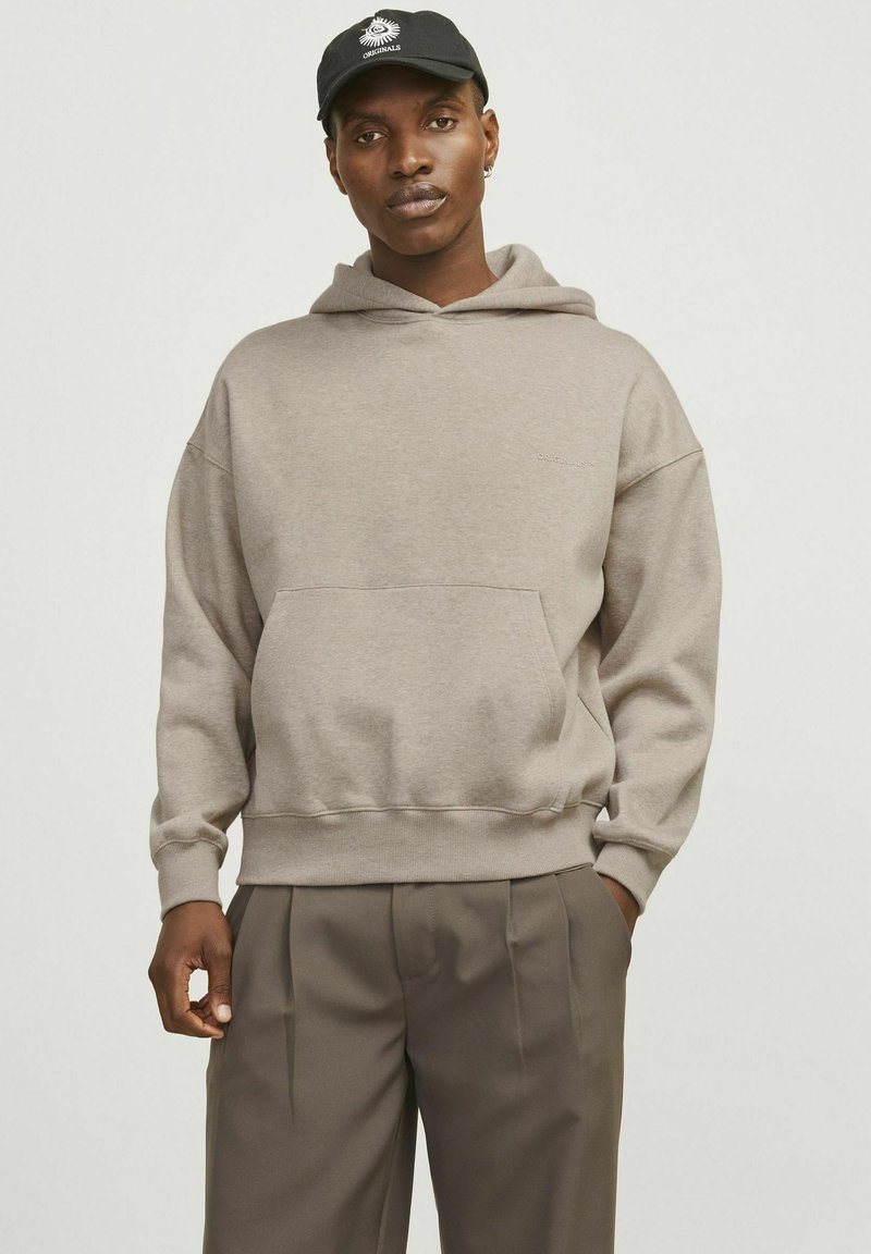 Beige hooded sweatshirt made of soft fabric, featuring a front pocket and ribbed cuffs, worn with tailored brown trousers.