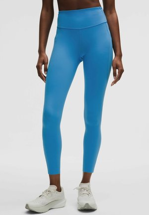 Frau, die hoch taillierte, leuchtend blaue Leggings und weiße Sportschuhe trägt, steht vor einem weißen Hintergrund.
