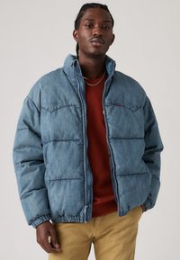 Denim pufferjakke, lys blå, oversized pasform; har en høj krave, elastiske manchetter og syninger; lagdelt over en rød sweater.