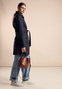 Granatowy płaszcz trench z paskiem, z detalami guzikowymi, zestawiony z jasnoniebieskimi spodniami typu wide-leg oraz białymi trampkami. Brązowa skórzana torebka na ramię.