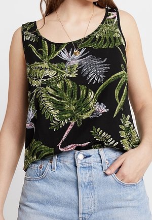 Vrouw draagt een zwarte mouwloze top met groene tropische bladeren- en bloemenprint, gecombineerd met lichtblauwe jeans met hoge taille.