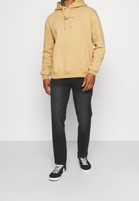Hombre vestido con una sudadera beige y jeans negros, usando zapatillas en blanco y negro, de pie frente a un fondo blanco liso.