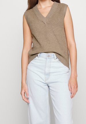 Frau trägt eine ärmellose braune Strickweste und hoch taillierte hellblaue Jeans, steht vor einem schlichten Hintergrund.