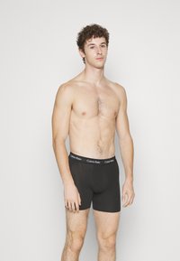 Zwarte katoenen boxershorts met een gladde textuur, voorzien van een merkband. Het model staat met een neutrale uitdrukking tegen een effen achtergrond.