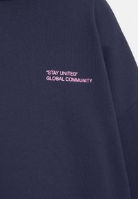 Indumento blu scuro con testo rosa "STAY UNITED" "COMUNITÀ GLOBALE" ricamato sul tessuto.