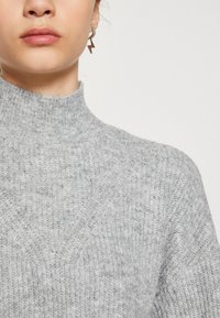 Sweater de malha cinza-claro com colarinho alto, apresentando um padrão texturizado. Acompanhado de um brinco geométrico com um design de raio.