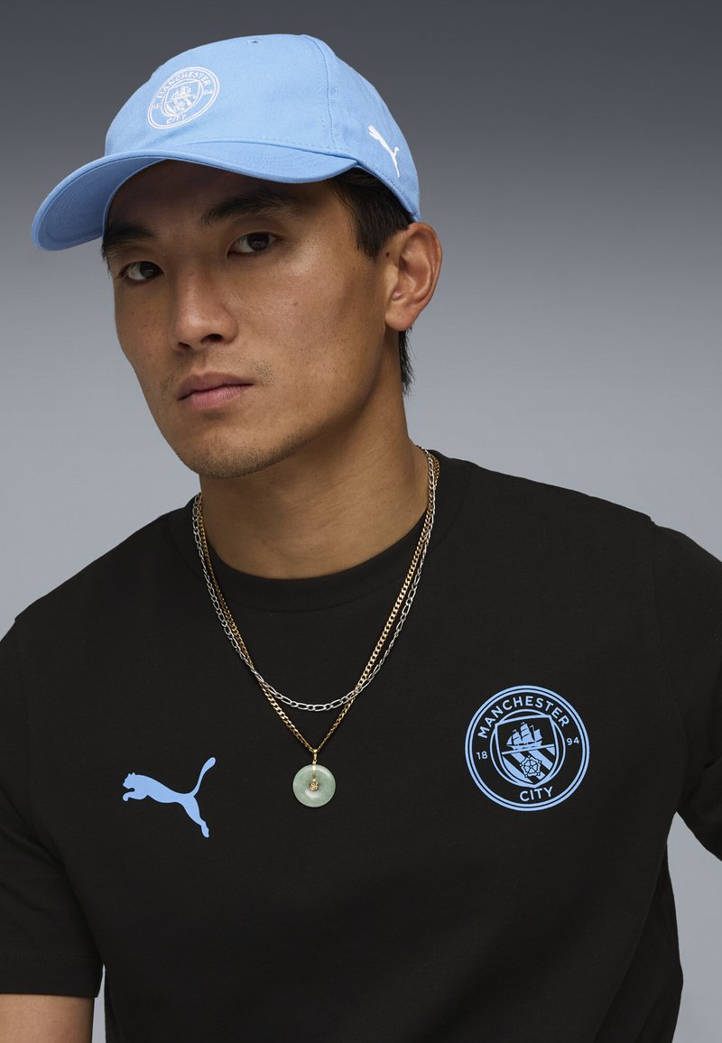 Homme portant une casquette Manchester City bleu clair et un t-shirt noir avec les logos Manchester City et Puma, deux chaînes autour du cou, fond gris neutre.