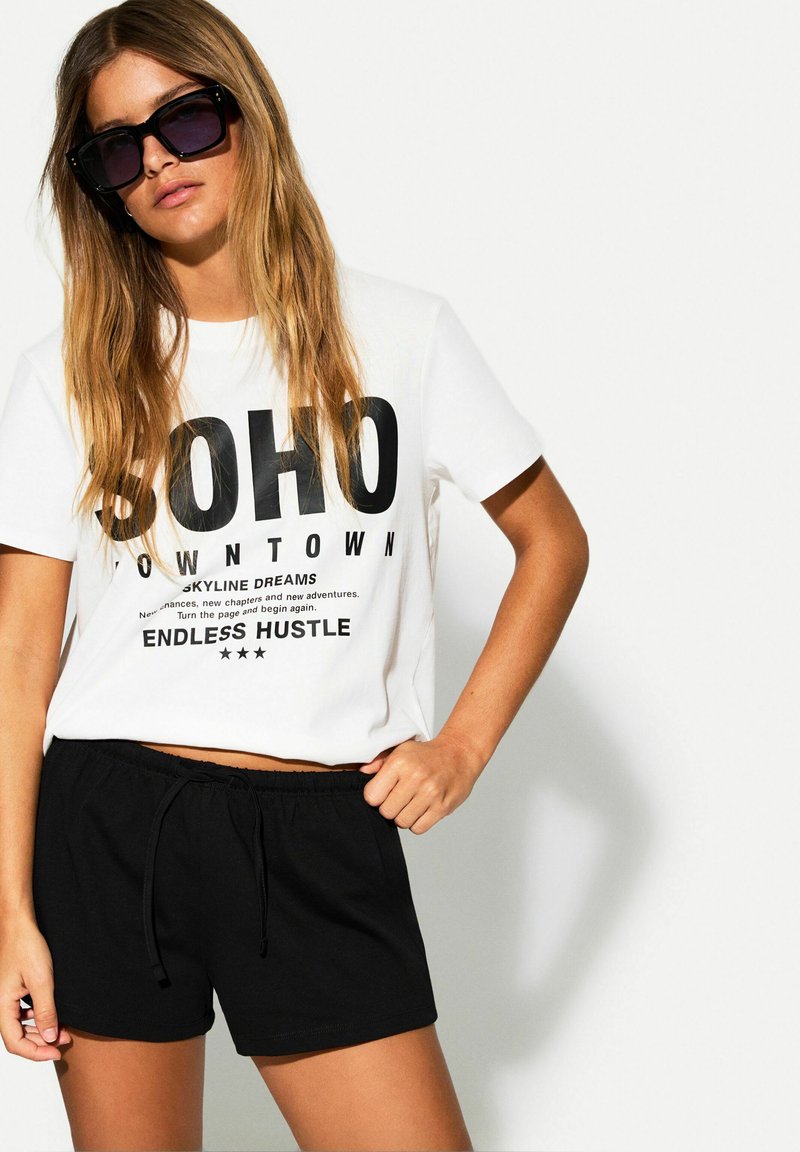 Biely tričko s krátkym rukávom s čiernym textovým grafickým motívom, skombinované s čiernymi šortkami na šnúrku. Model nosí oversized čierne slnečné okuliare.