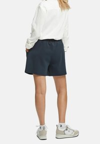 Mörkblå shorts med elastisk midja, avslappnad passform och sidofickor, kombinerat med en vit skjorta med knappar och grå sneakers.