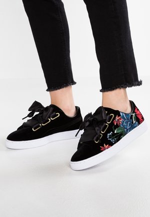 Schwarze Samtsneaker mit großen Satin-Schleifen und bunter Blumenstickerei, getragen zu schwarzen Jeans mit ausgefranstem Saum auf weißem Boden.