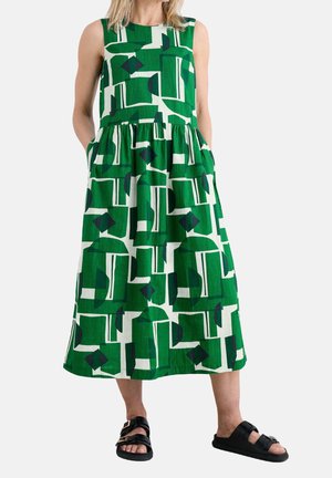 Robe midi sans manches à motif géométrique abstrait vert et blanc, portée avec des sandales noires à boucle, mains dans les poches latérales.