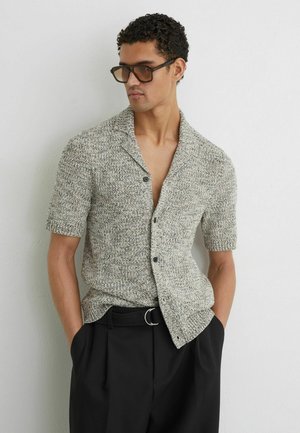 Mężczyzna w teksturowanym, krótkim, zapinanym na guziki kardiganie, czarnych plisowanych spodniach z paskiem w kształcie pierścienia, dużych brązowych okularach przeciwsłonecznych, z rękami w kieszeniach.