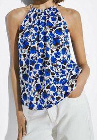 Top a fascia floreale con fiori blu e neri su uno sfondo bianco. Tessuto leggero, scollo arricciato, vestibilità morbida. Indossato con pantaloni bianchi.