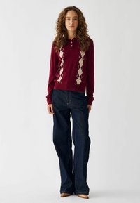 Suéter de polo burdeos con patrón de rombos crema, combinado con vaqueros de pierna ancha azul oscuro. El suéter tiene un cuello y tres botones en el cuello.