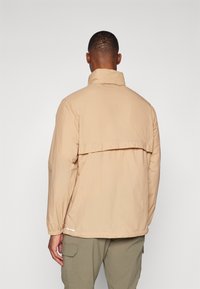 Veste légère beige avec un col montant, une coupe décontractée, des poignets élastiques et un ourlet allongé à l'arrière. Présente des détails de couture subtils et des accents blancs.