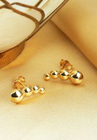 Orecchini in oro con un design graduato di sfere lisce e riflettenti di varie dimensioni, su uno sfondo morbido e beige.
