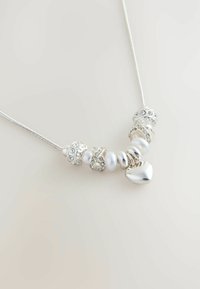 Hõbedane kaelakee, millel on mitmesuguseid helmeid, sealhulgas poleeritud valged pärlid ja säravad kristallaktsendid, südamekujulise ripatsiga.