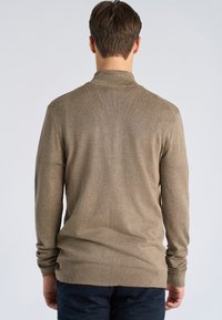 Homme debout de dos, portant un pull à manches longues marron clair et un pantalon bleu foncé, sur un fond uni clair.