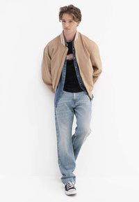 Braune Bomberjacke mit weiß gestreiftem Kragen über einem Jeanshemd und schwarzem T-Shirt; hellblaue Jeans und schwarz-weiße Turnschuhe.