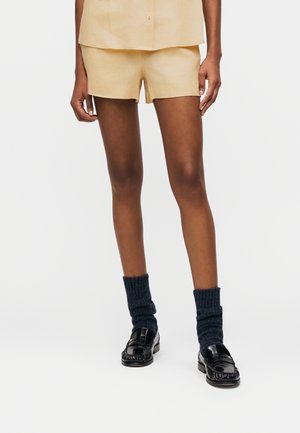 Persona vestida con pantalones cortos beige y top a juego, combinados con calcetas oscuras de punto y mocasines de cuero negros sobre un fondo liso.