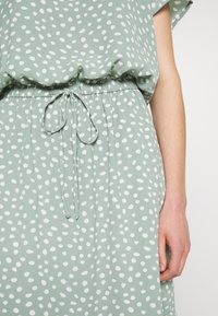 Robe vert clair avec des pois blancs irréguliers, taille élastique nouée à l'avant, et un bras droit visible reposant à côté du corps.
