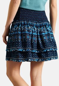 Blå mönstrad kjol med en smockad midja, lagerdesign och intrikata blommiga och prickiga mönster mot en mörk bakgrund.