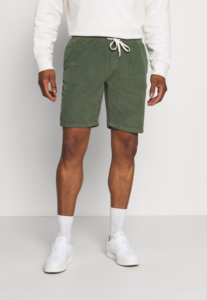 Grüne Cordshorts mit einem elastischen Bund, Kordelzug und Seitentaschen, kombiniert mit weißen Socken und Turnschuhen.
