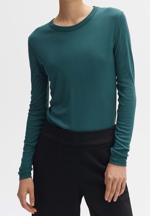 Personne portant une chemise ajustée à manches longues couleur sarcelle rentrée dans un pantalon noir taille haute sur un fond uni.
