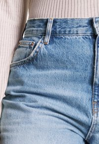 Jeans de mezclilla azul de talle alto con un lavado claro, que cuentan con un bolsillo delantero y costuras en color contrastante a lo largo de las costuras. Textura ligeramente desgastada.