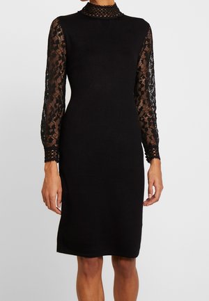 Robe noire longueur genou avec manches longues en dentelle florale transparente et col montant en dentelle portée par une personne sur fond uni.