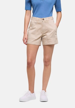 Mujer con shorts beige de talle alto con bolsillos frontales, camiseta azul de manga corta y zapatillas blancas, de pie con las manos en los bolsillos.