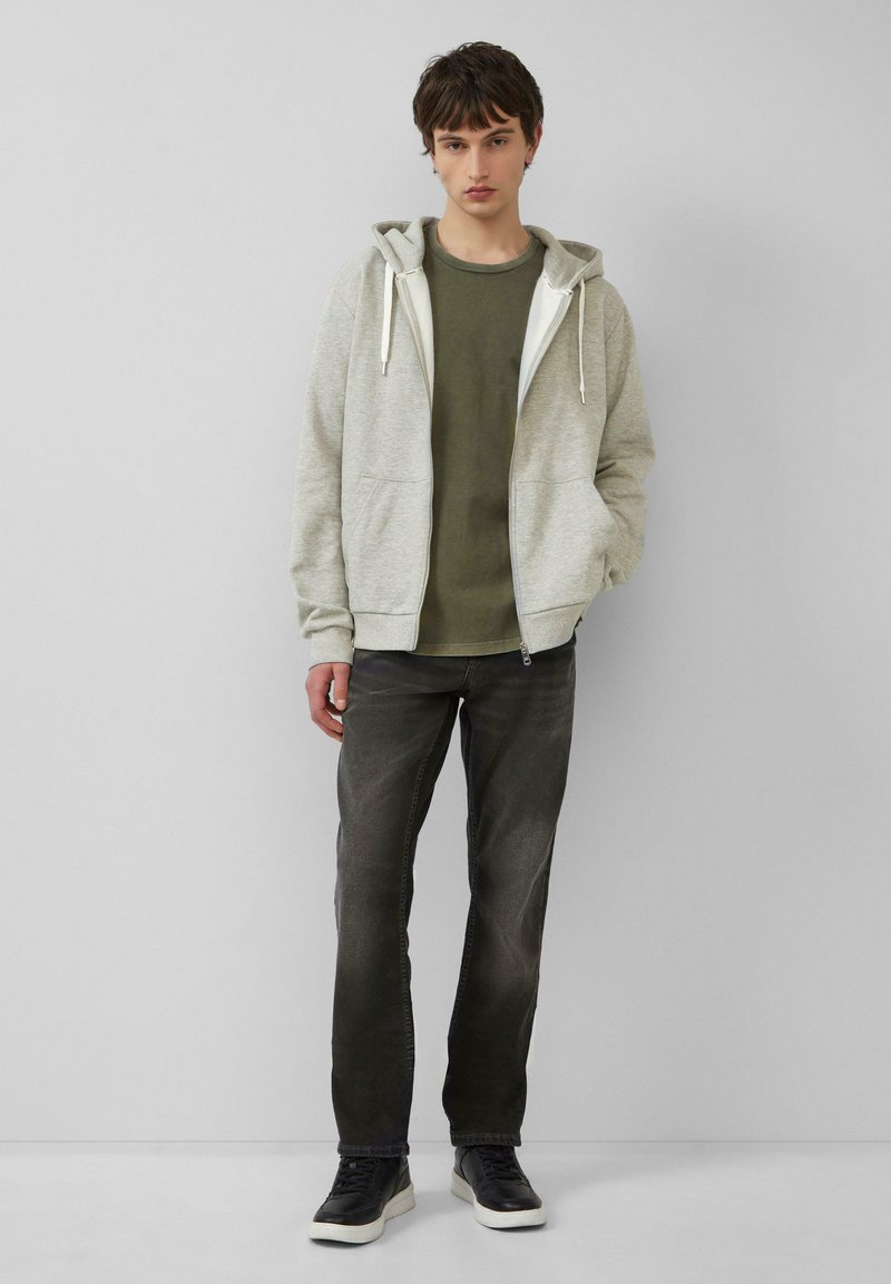 Jasnoszara bluza z kapturem na suwak z białym podbiciem, nałożona na oliwkową koszulkę, w zestawieniu z ciemnoszarymi dżinsami i czarnymi trampkami.