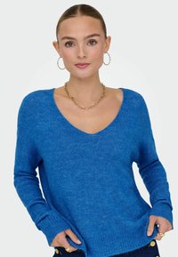Femme portant un pull à col en V bleu vif, boucles d'oreilles créoles en or, collier chaîne et bagues, posant les mains dans les poches devant un fond uni.