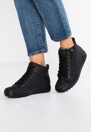 Pieds portant des baskets montantes noires avec une fermeture éclair latérale et des lacets épais, assorties à un jean bleu retroussé sur un sol blanc.