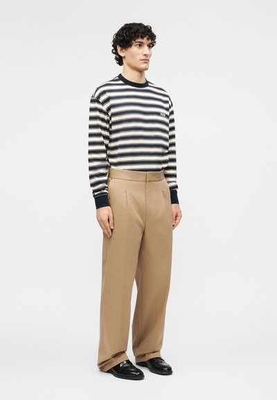 Άνδρας που στέκεται φορώντας πουκάμισο με μακριά μανίκια σε χρώματα ναυτικό μπλε, λευκό και μπεζ ριγέ, μπεζ παντελόνια φαρδιάς γραμμής και μαύρα δερμάτινα παπούτσια.