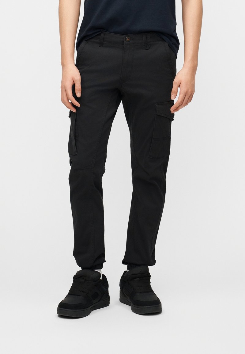 Homme portant un pantalon cargo noir avec des poches à rabat et des baskets noires, debout devant un fond clair uni.