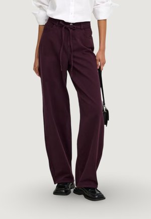 Persona che indossa pantaloni larghi viola scuro con vita legata, scarpe nere e una camicia bianca a maniche lunghe, che tiene una piccola borsa nera.